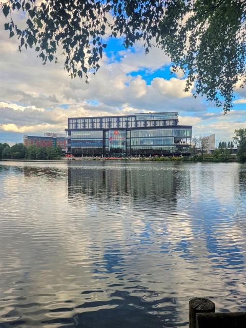 The Resorts World Birmingham complex seen across Pendigo Lake beneath a cloudy sky, with trees framing the view.