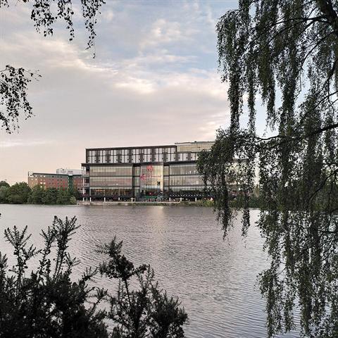 Evening view of the Resorts World Birmingham building across still water, with willow branches hanging into the frame.